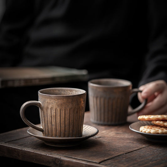 Two rustic ceramic cups on a wooden table with a person holding one of them.