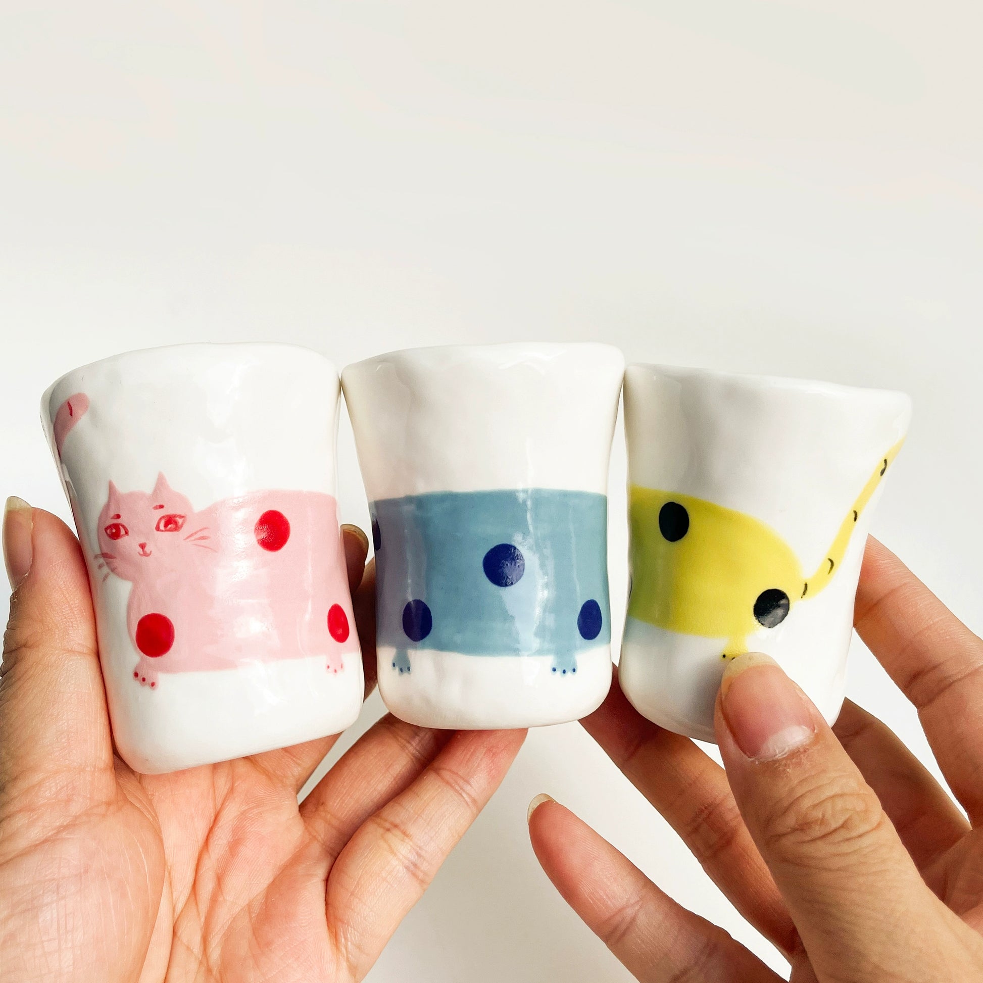 Three small ceramic cups with cat designs held by a hand against a white background